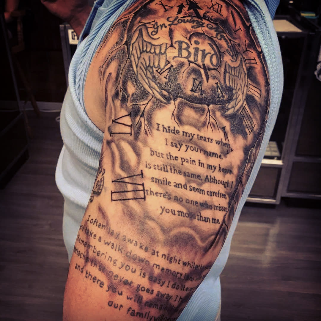 A detailed black and gray arm tattoo featuring wings, clocks, and an intricate poem